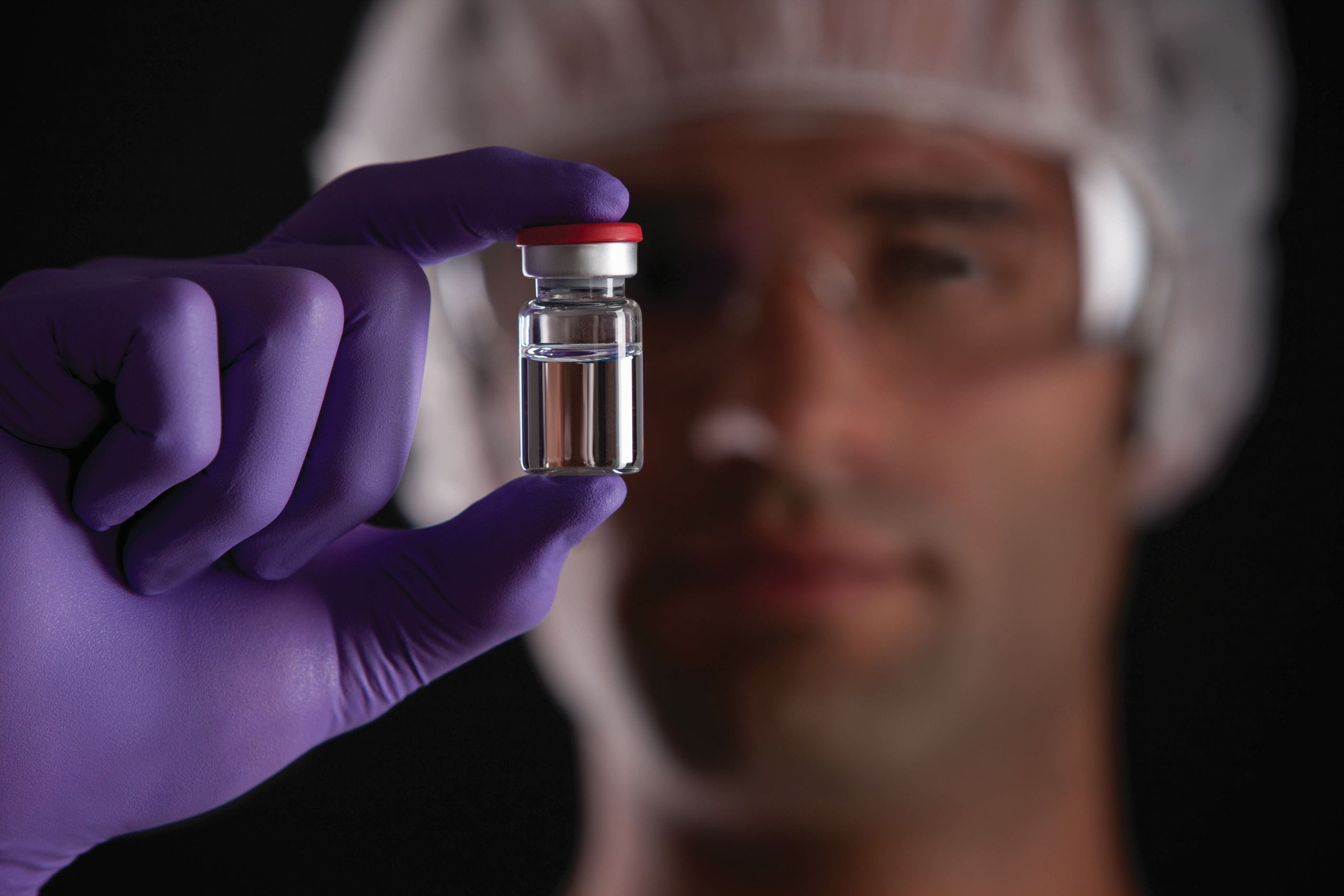 Gloved hand holding a sealed vial containing injectable liquid in a sterile environment.