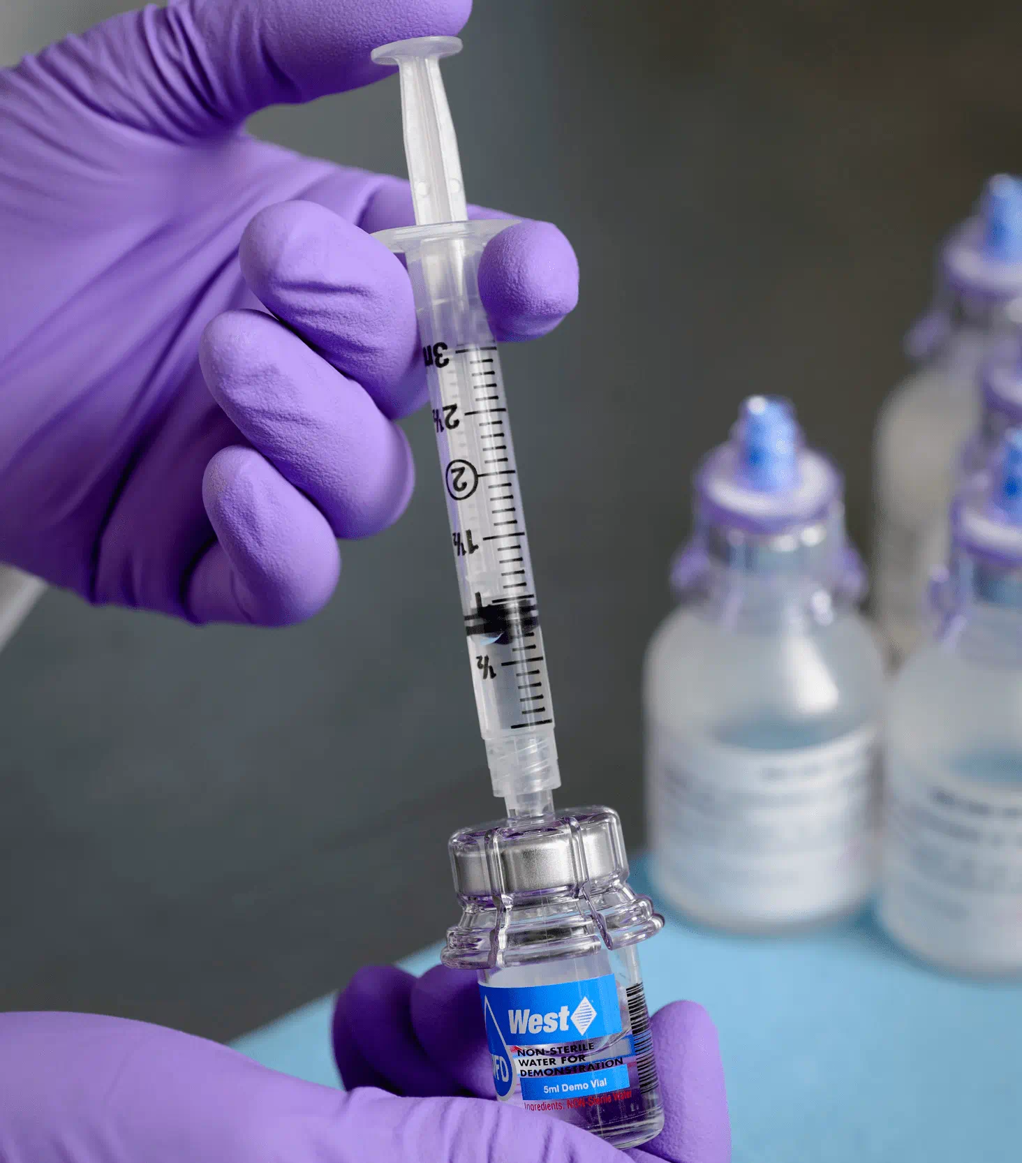Gloved hand drawing liquid into a syringe from a vial fitted with a medical adapter.