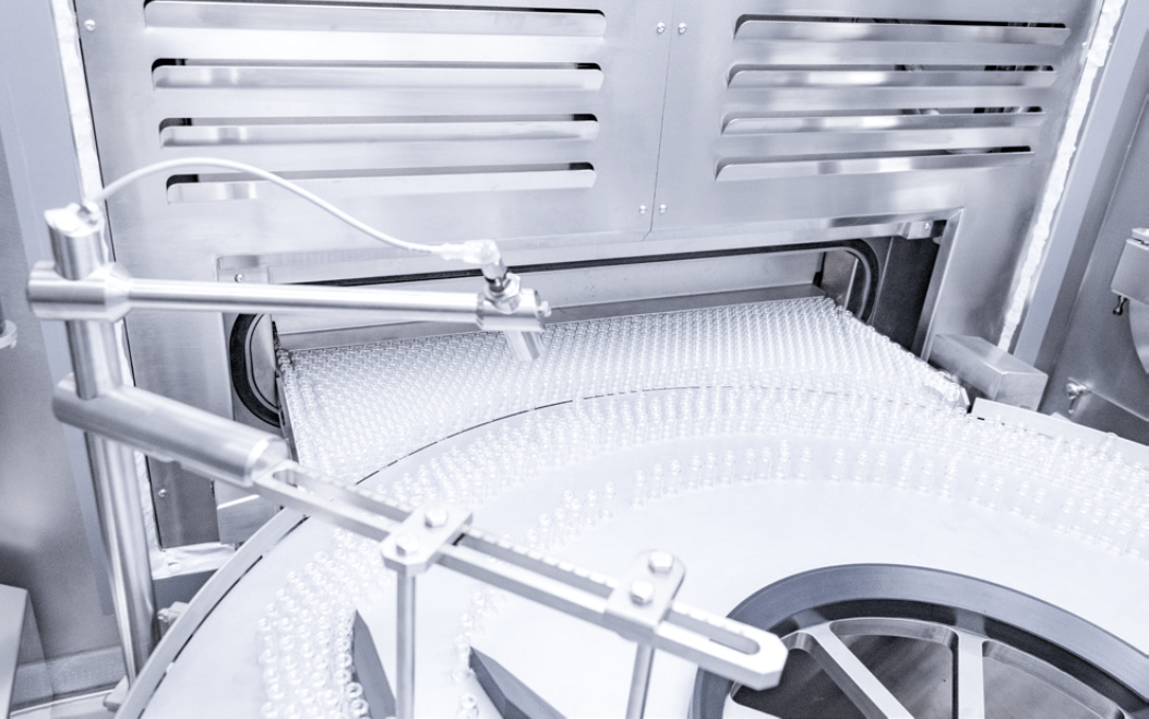 Aseptic pharmaceutical production line with trays of glass vials moving through automated sterile processing equipment.