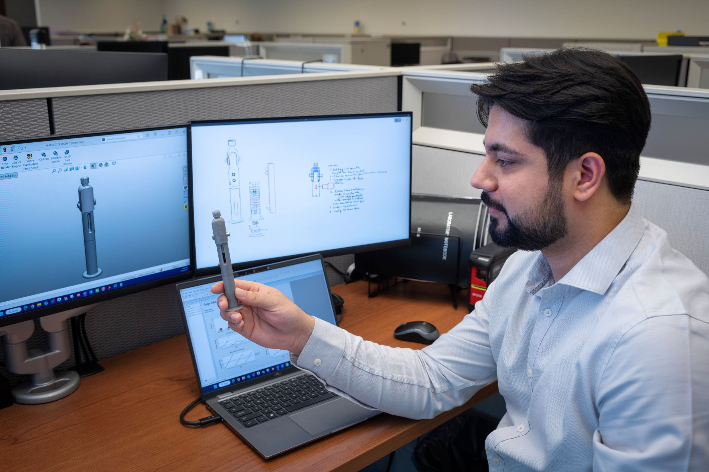 Engineer reviewing a 3D printed component alongside CAD design models on dual monitors.