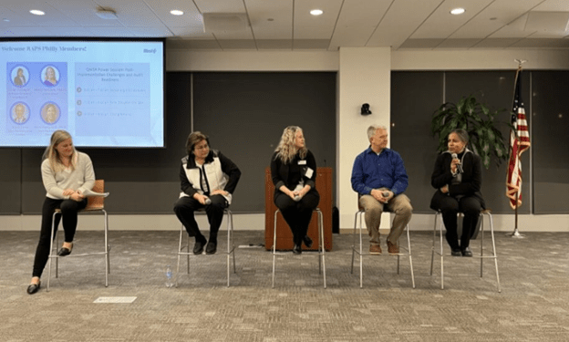 Panel of speakers seated onstage during the RAPS Philadelphia QMSR Power Session with a presentation screen on the left and a U.S. flag on the right